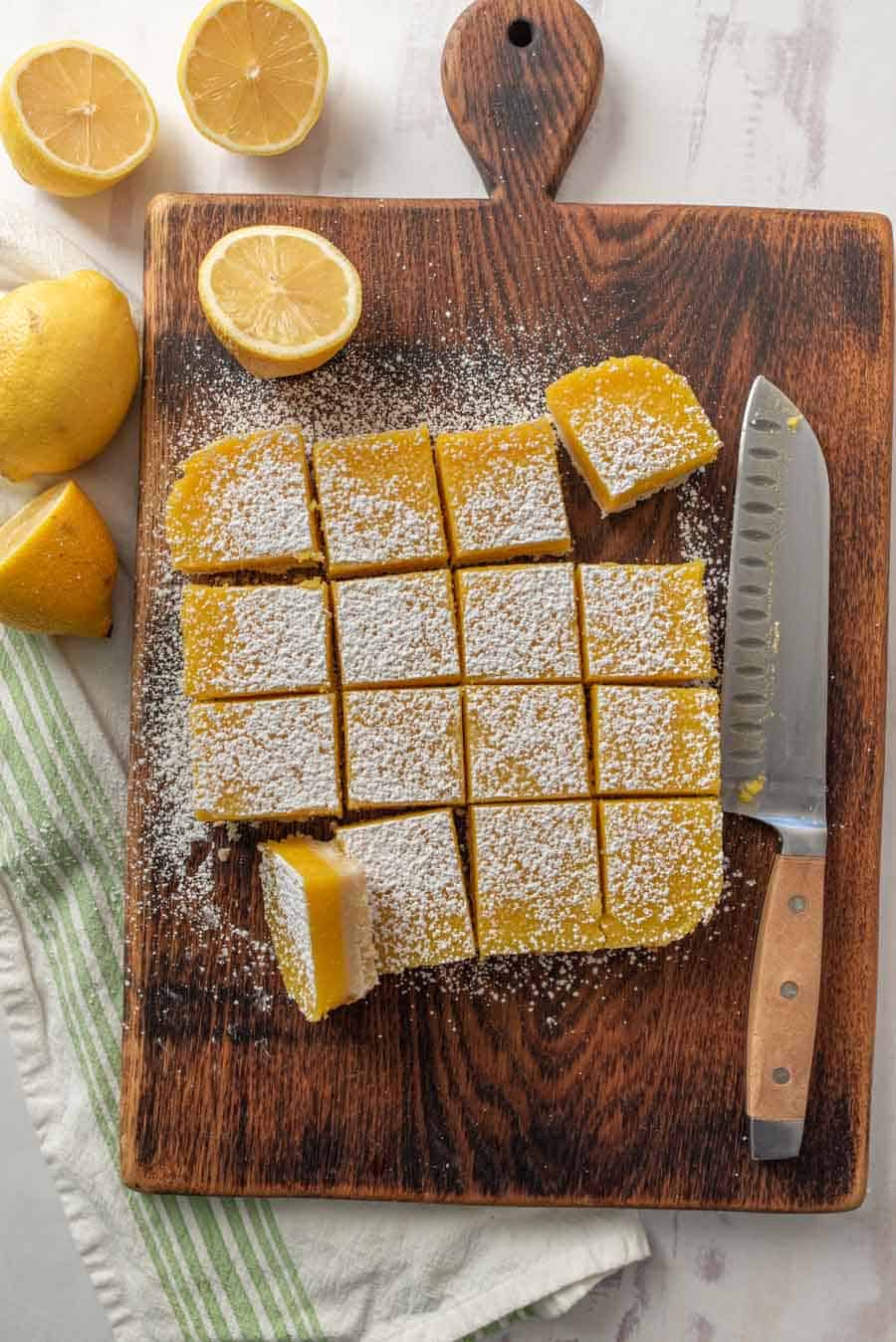 Top view of lemon bars on wood cutting board with knife and lemons around it.