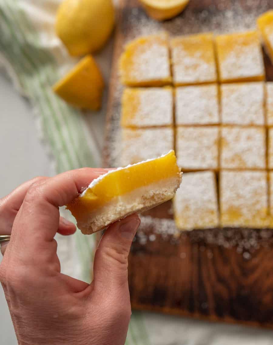 Hand holding a lemon bar with the rest of the pan of cut lemon bars in the background.