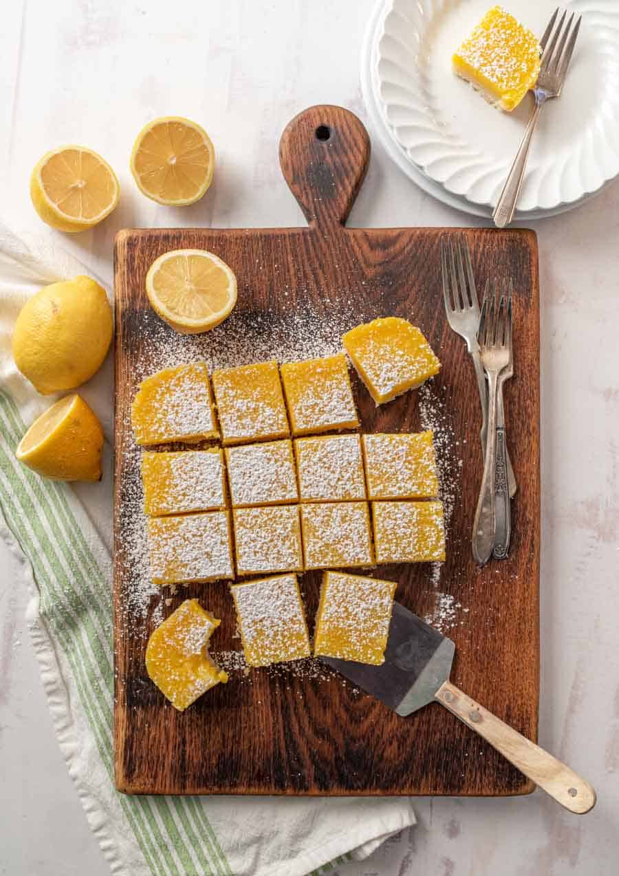 Top-down view of lemon bars dusted with powdered sugar, cut into squares on a wooden board with a spatula, surrounded by fresh lemons and forks, with a plate of lemon bars in the upper right.