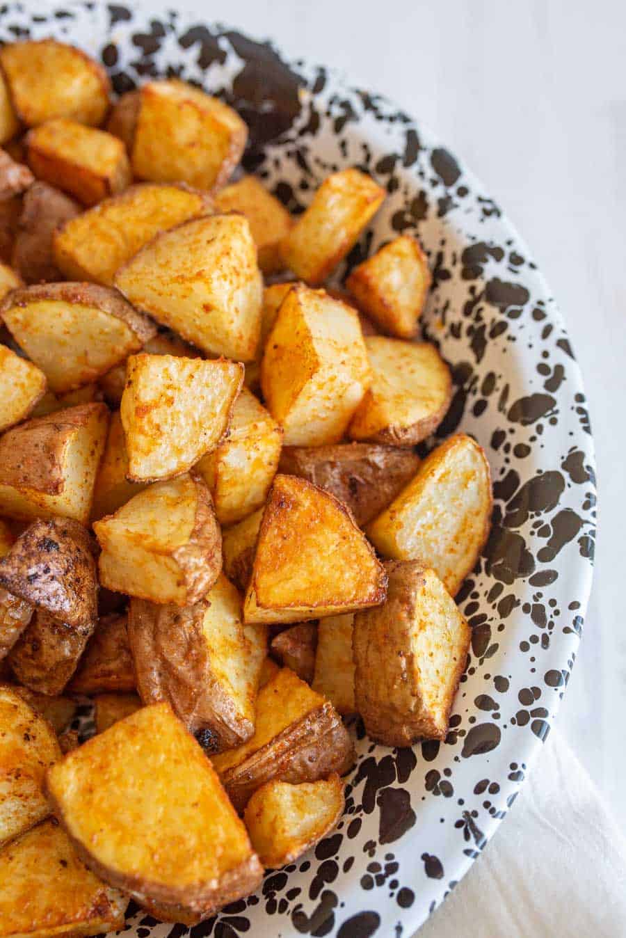 A close-up of a plate filled with oven roasted red potatoes, golden brown with seasoning, served on a white dish with black speckles.
