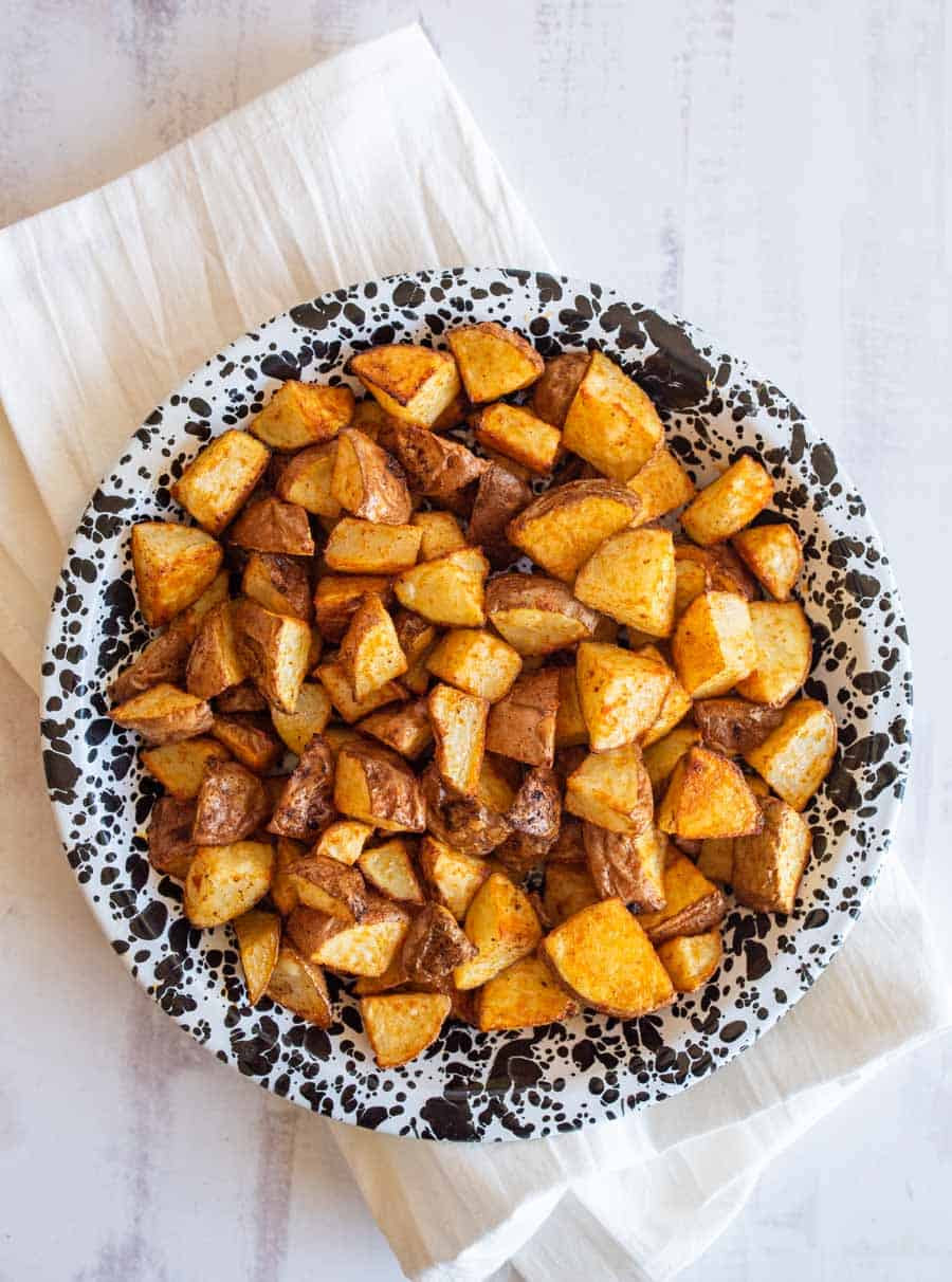 A plate filled with golden oven roasted red potatoes, served on a black and white speckled plate placed on a cream-colored cloth. The potatoes have a crispy, seasoned appearance.