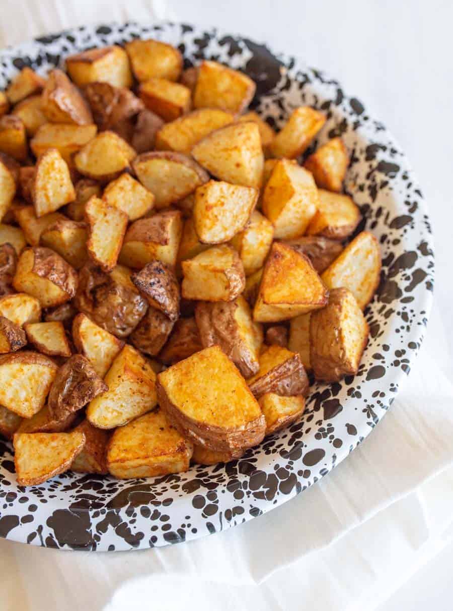 A plate filled with golden-brown, oven roasted red potatoes seasoned with spices, placed on a white cloth. The plate features a black and white splatter pattern design.