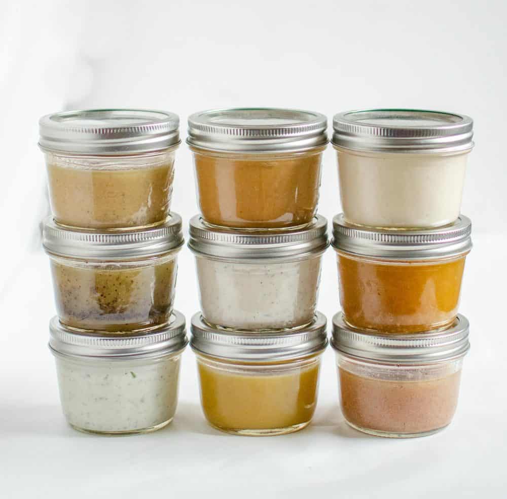 Nine small glass jars with metal lids are stacked in three rows, each containing different colored sauces or dressings that inspire new ideas for how to make salad dressing, all set against a plain white background.