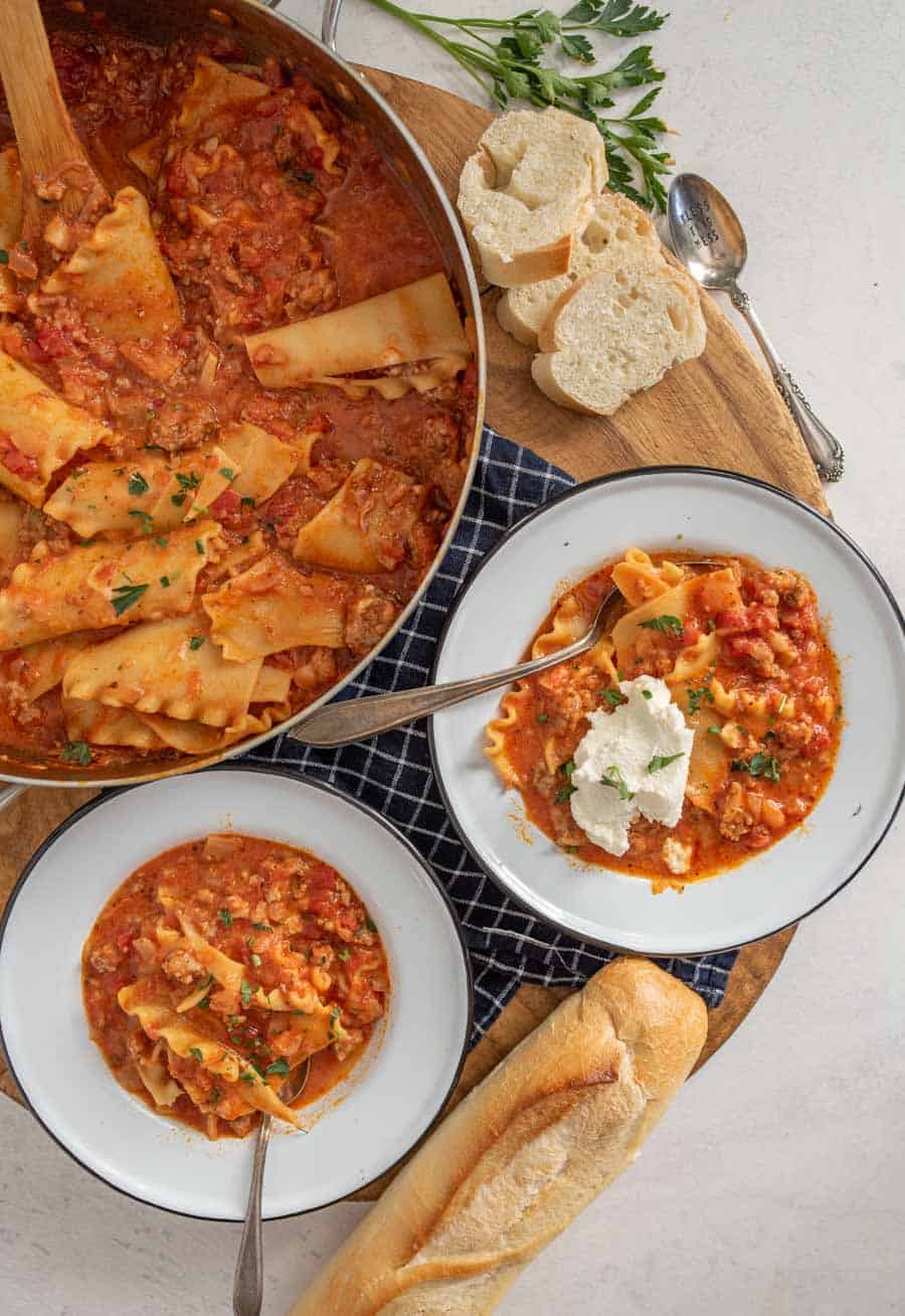 A top down view of lasagna soup in pan next to two bowls and a loaf of french bread.