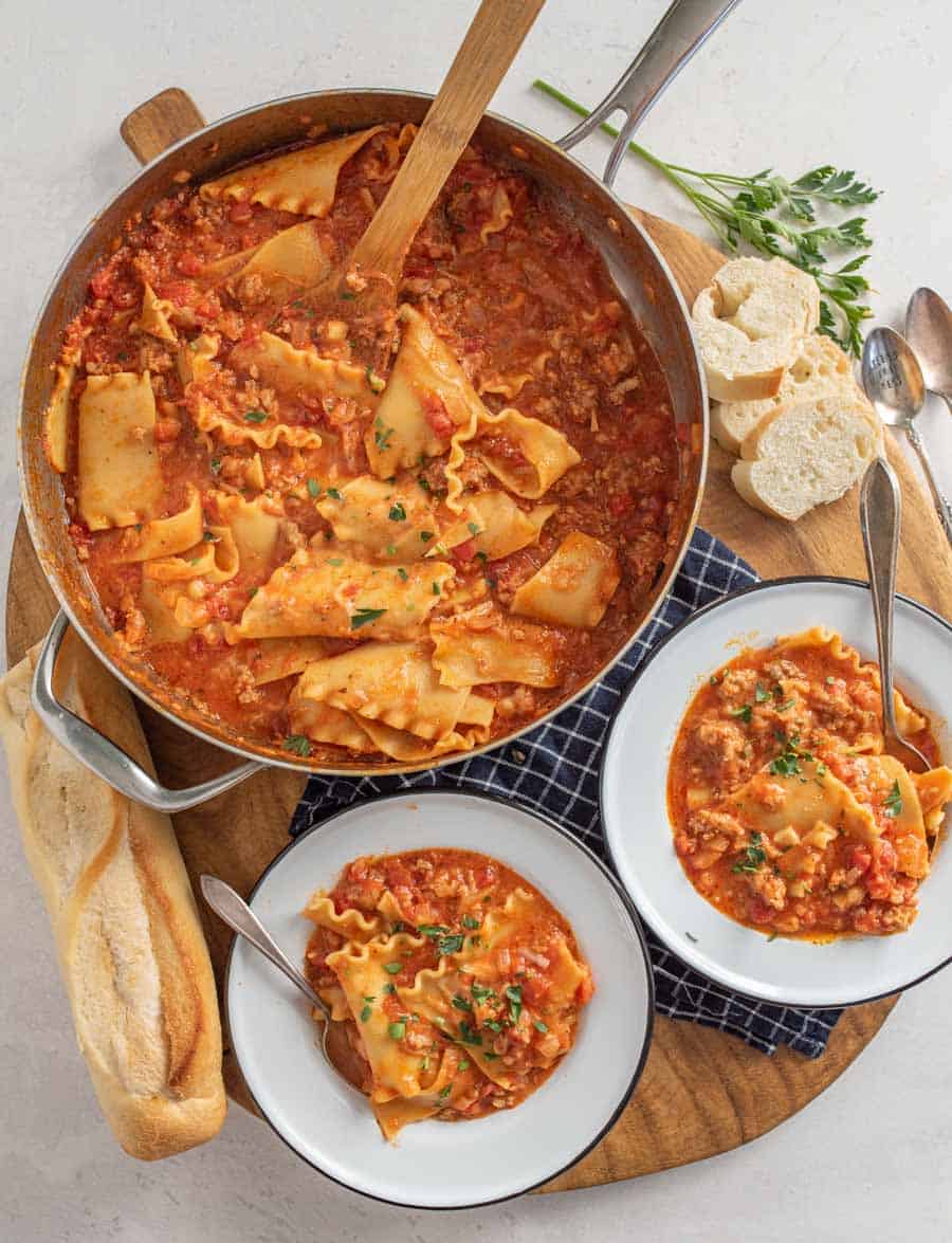 A top view of pot of lasagna soup with two white bowls full of the soup and a loaf of French bread.