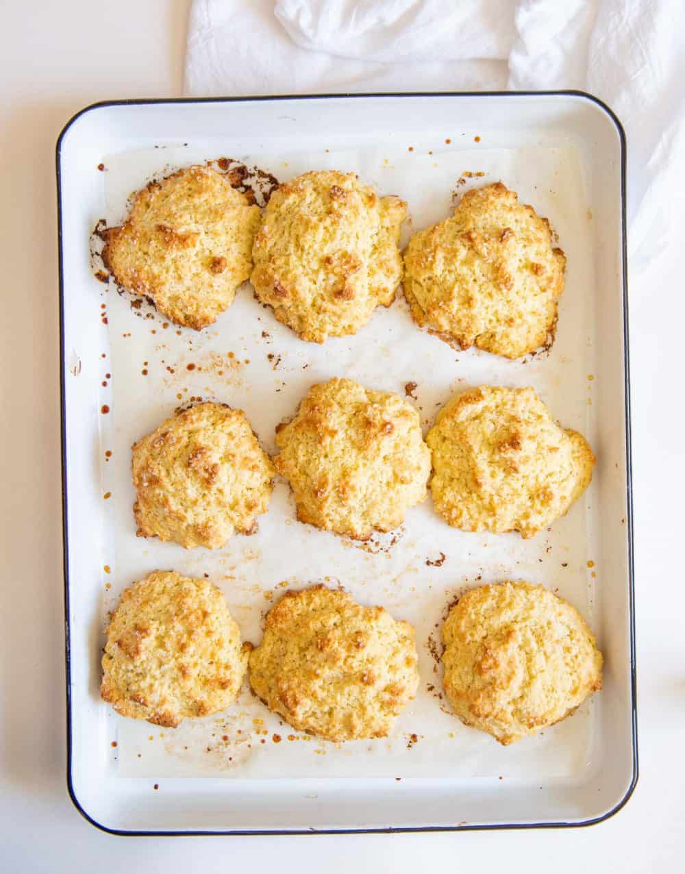 Eight golden-brown drop biscuits, perfect for a Biscuit Recipe for Strawberry Shortcake, are arranged on a parchment-lined white baking tray, fresh out of the oven. A white cloth is partially visible in the background.