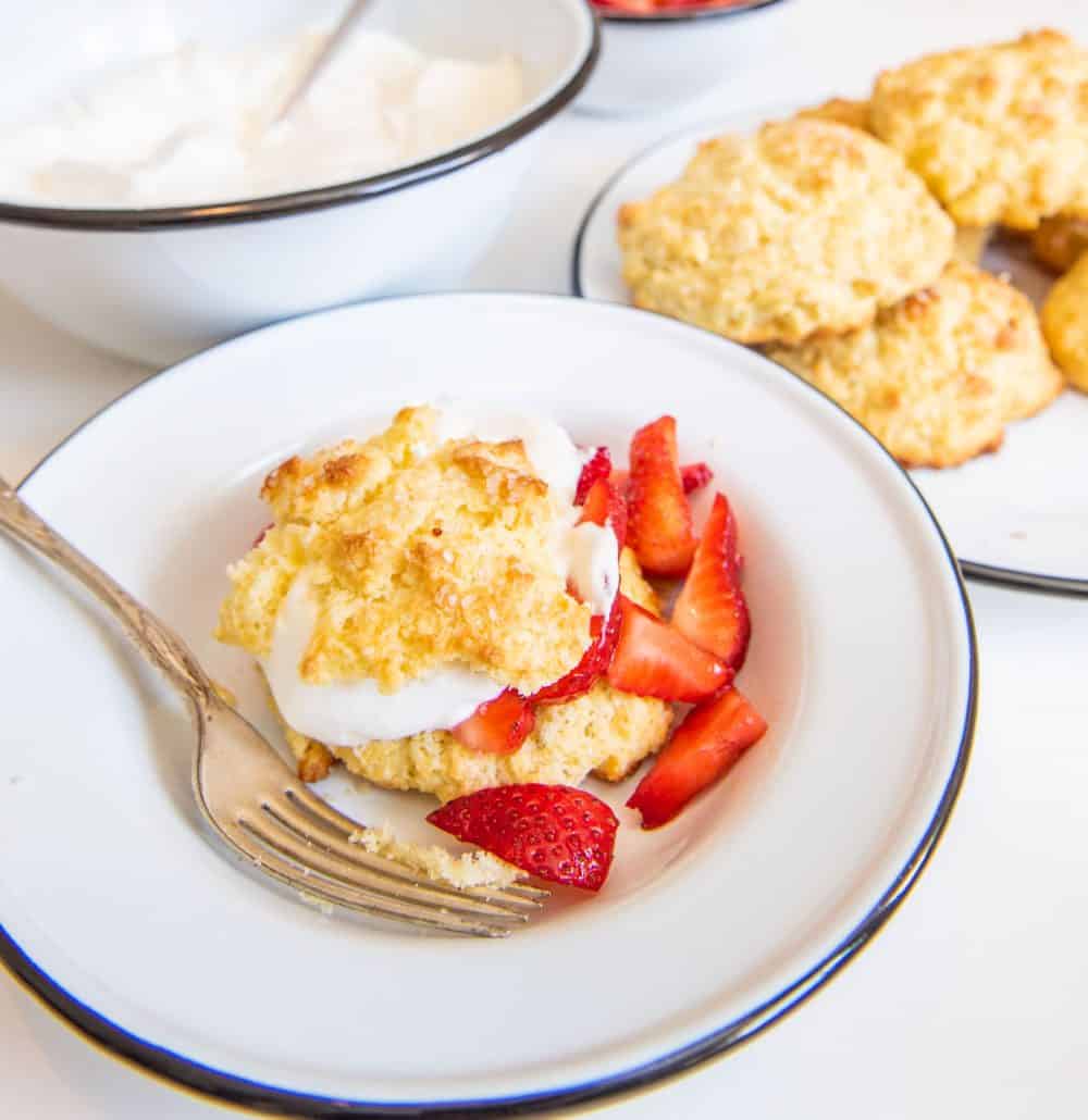 A bowl with a strawberry short cake biscuit, whipped cream, and sliced strawberries with a fork and short cake biscuits in the background.