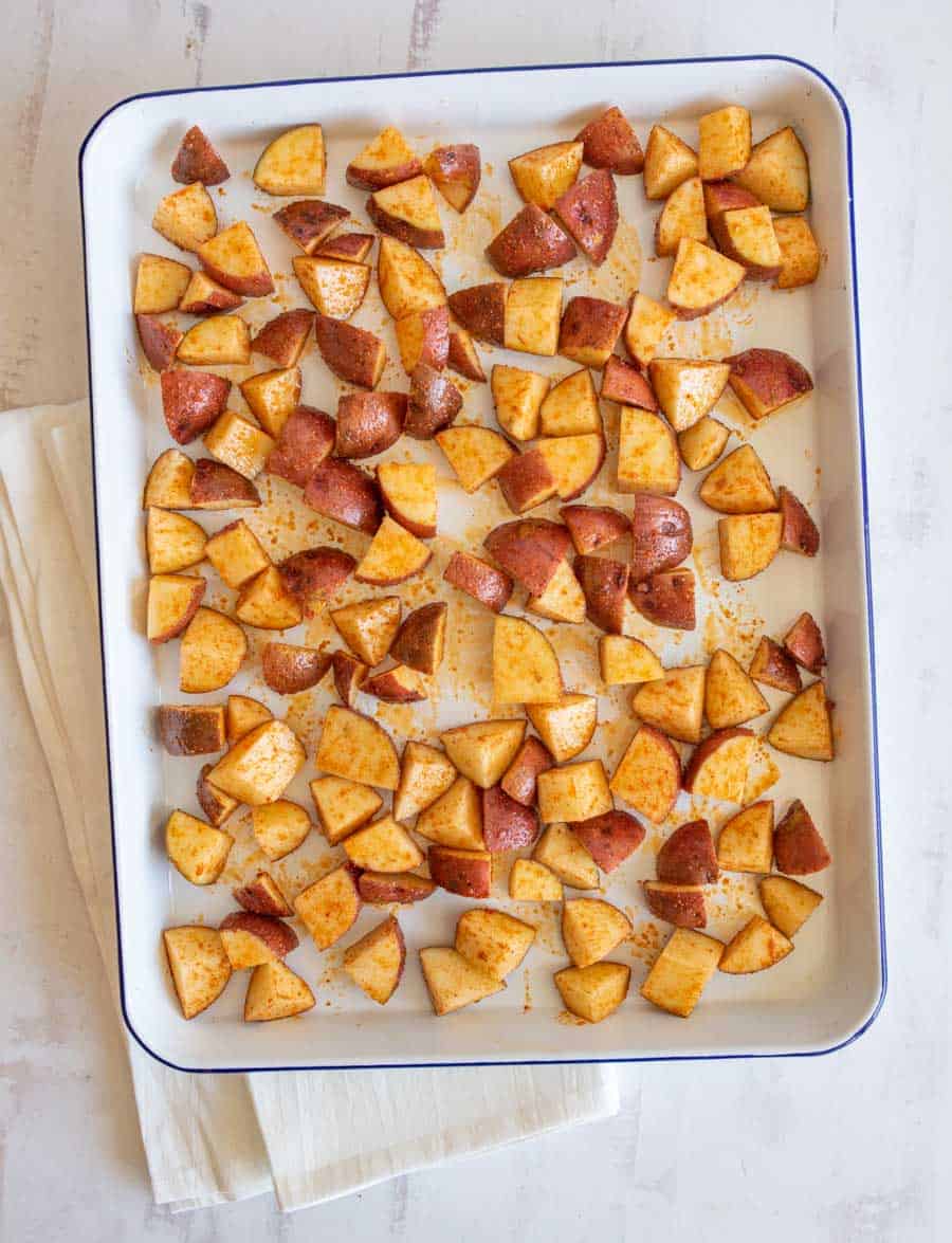 A white baking tray filled with evenly spread, seasoned, diced red potatoes, ready for oven roasted red potatoes. The tray rests on a folded cream-colored cloth on a light-colored surface.