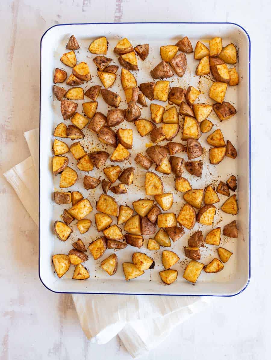 A baking sheet filled with evenly spread, golden-brown oven roasted red potatoes cubes on white parchment paper. The sheet rests on a white cloth over a light-colored surface.