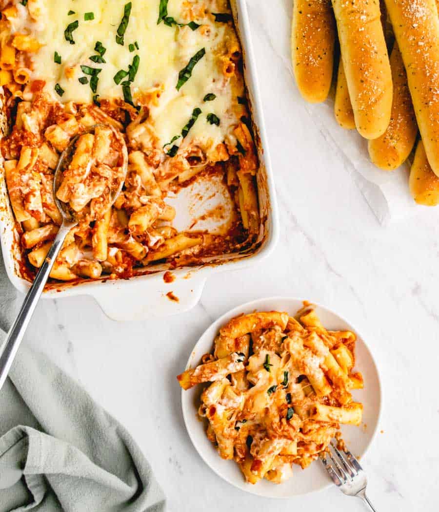 Top view of a plate and baking dish of baked ziti with breadsticks on the side.