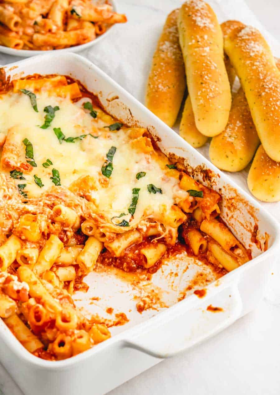 A white baking dish with baked ziti in it with breadsticks in the background.