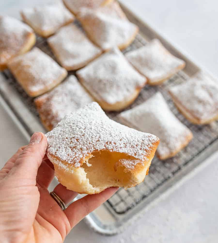 A hand holding a beignet that has a bite out of it with a tray of beignets in the background.