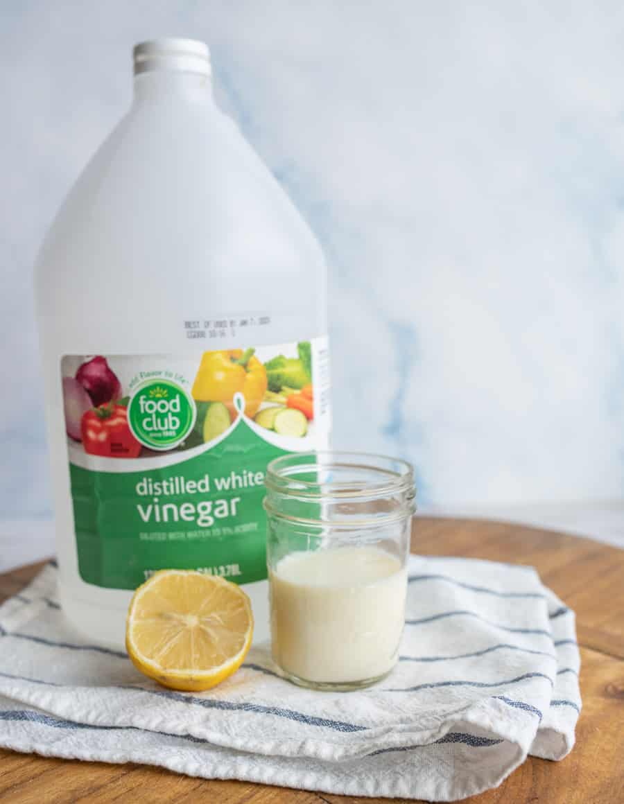 A jug of white vinegar, half a lemon, and a glass of milk on a white napkin on a wood cutting board.