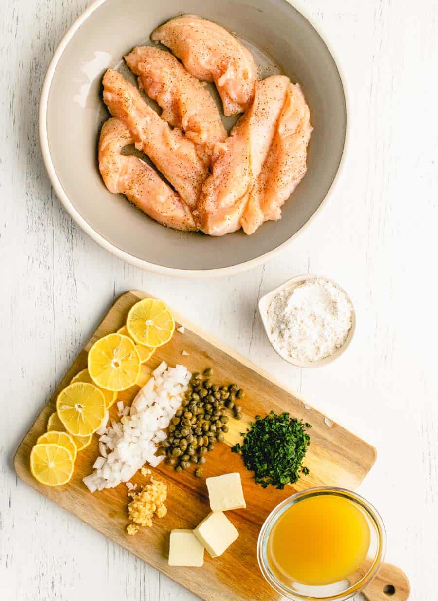 A top view of a bowl of chicken and a cutting board with the ingredients for chicken picatta on it.