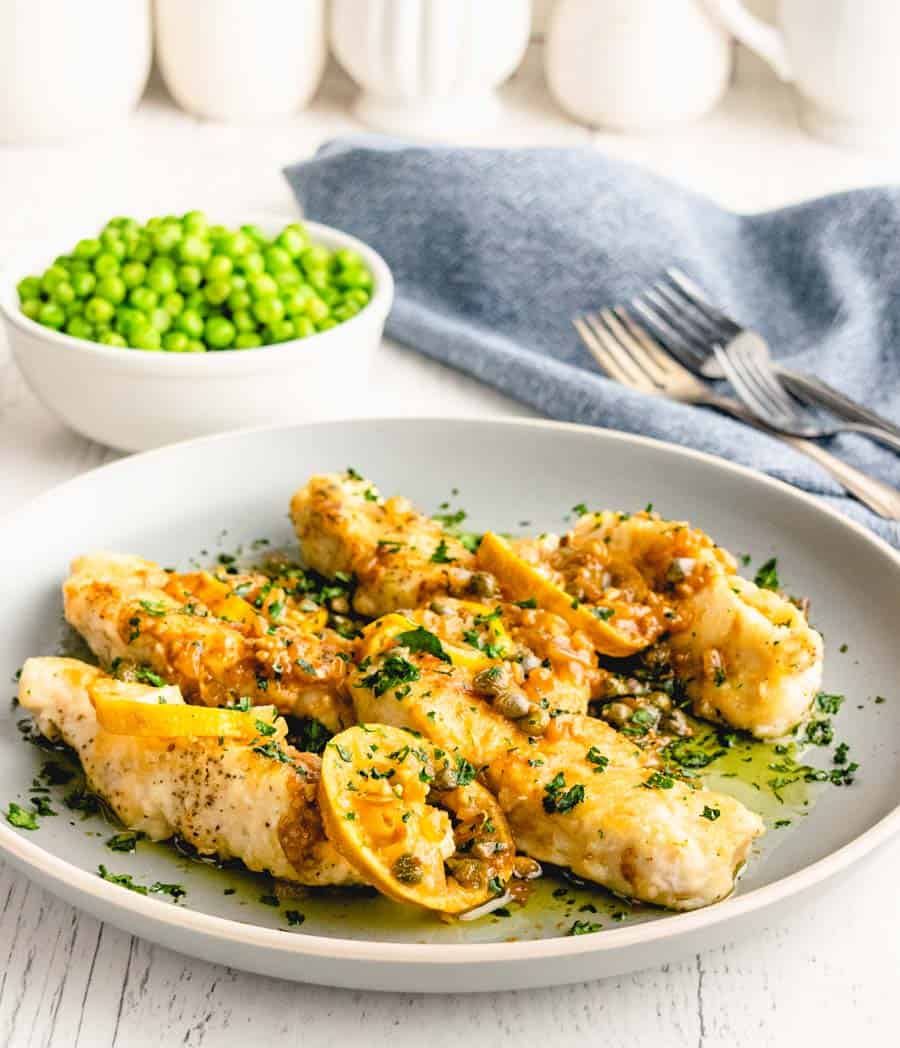 A plate of chicken picatta with a bowl of peas and forks in the background.