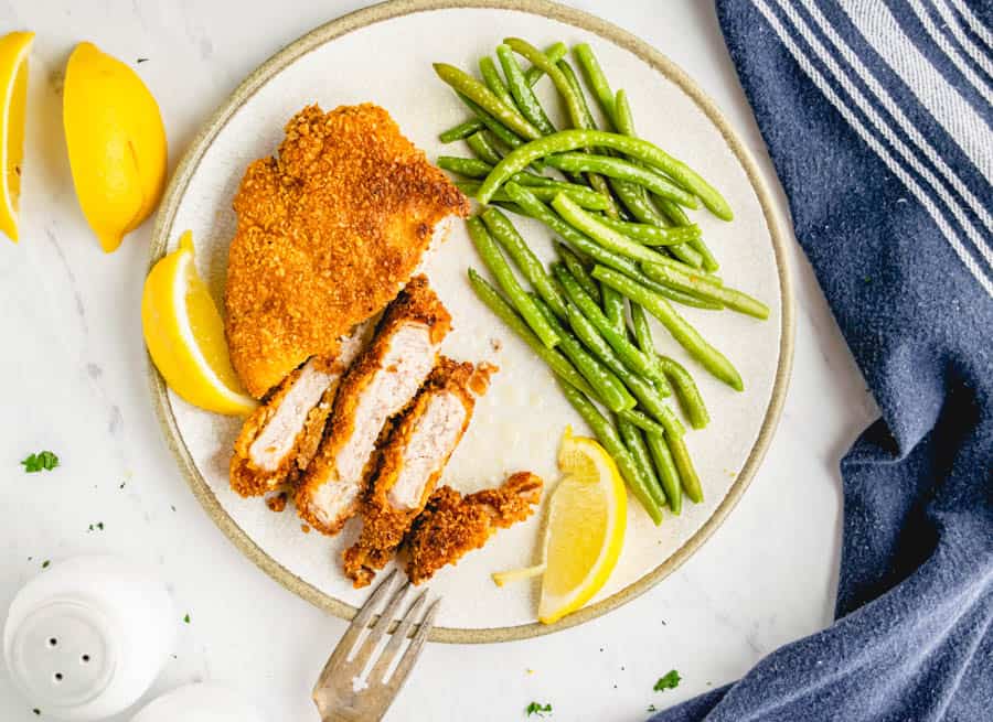 Top view of a plate with green beans and a crispy fried pork chop cut into slices with lemon slices.
