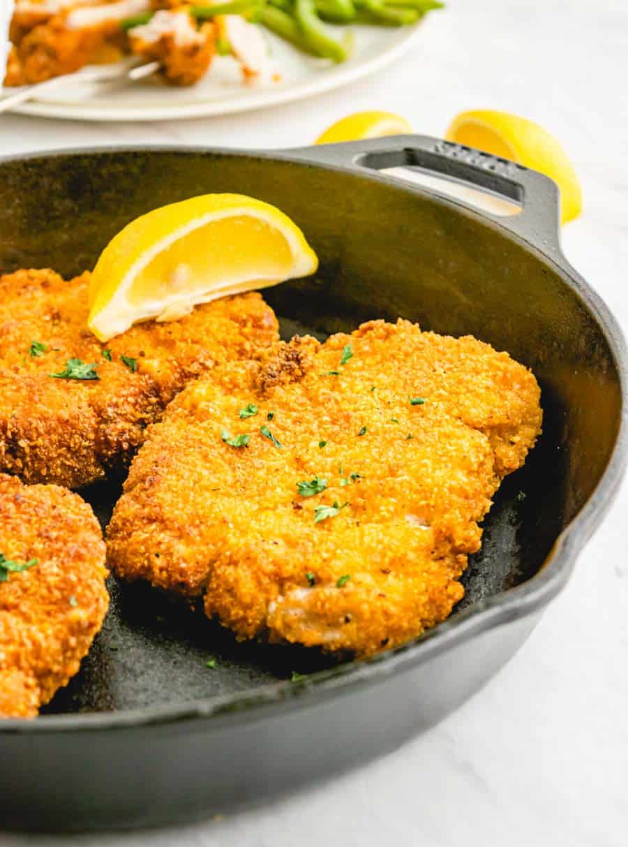 Close up picture of crispy fried pork chops in a cast iron skillet with a lemon slice on top of one.