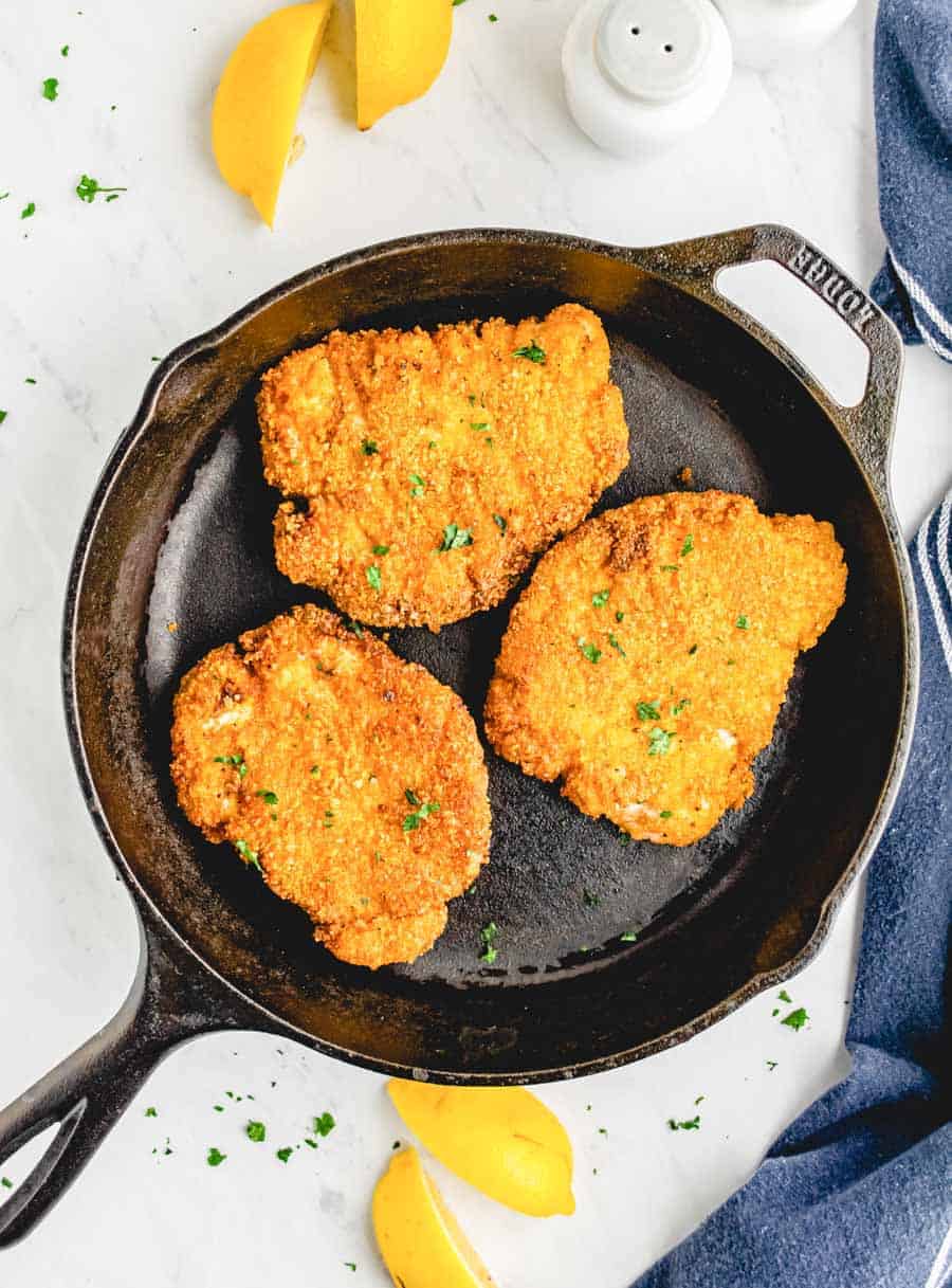 Crispy fried pork chops in a cast iron skillet with lemon wedges next to it.