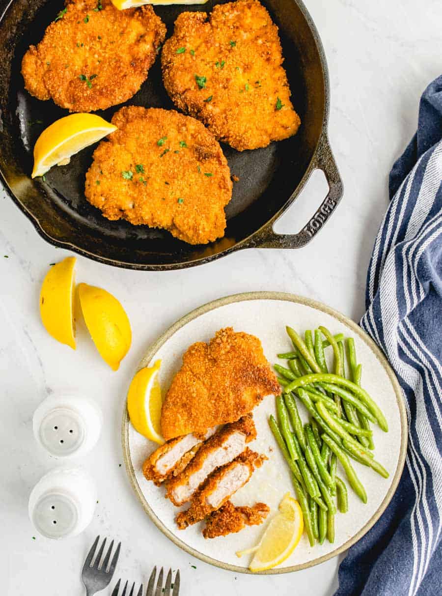 Top view of crispy fried pork chops in a cast iron skillet next to a plate of sliced pork chop and green beans.