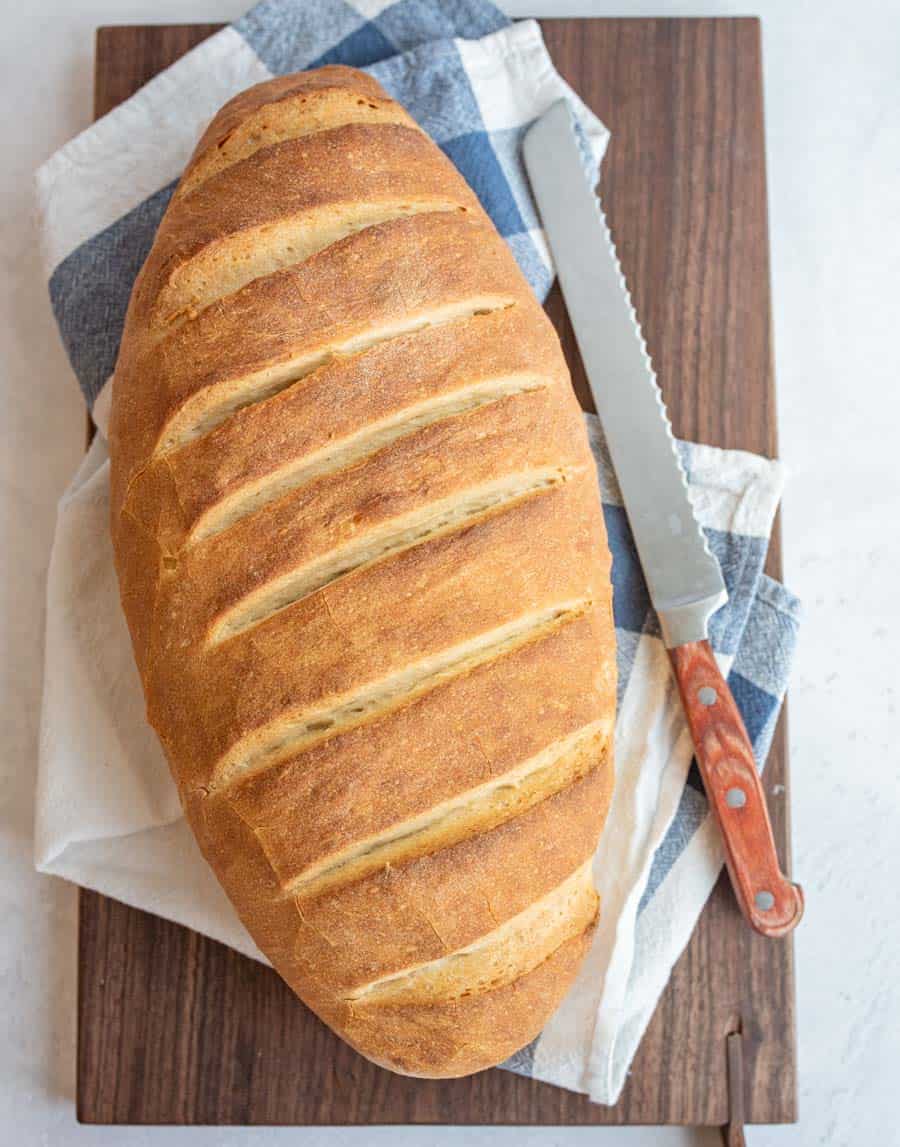 Top view of a loaf of Italian bread on a towel and wooden cutting board with a knife next to it.