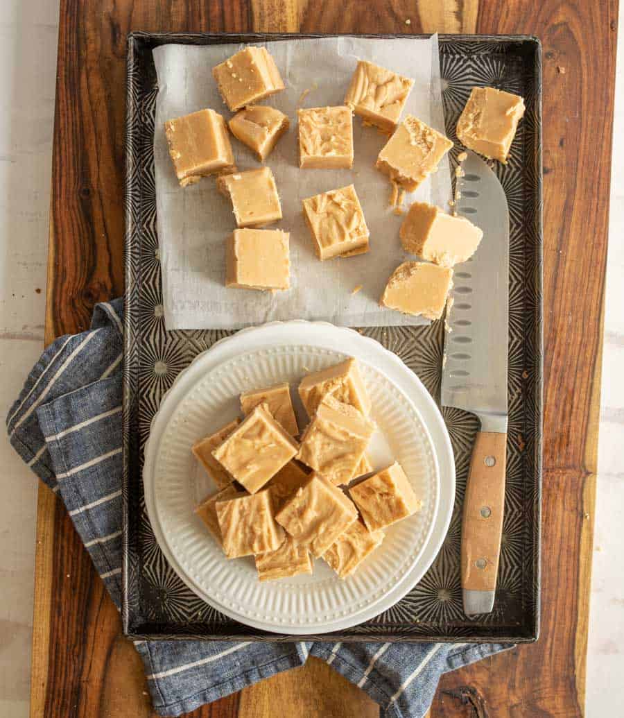 Top view of a pan with squares of peanut butter fudge on parchment paper, and some on a stacked on a white plate with a knife next to it.
