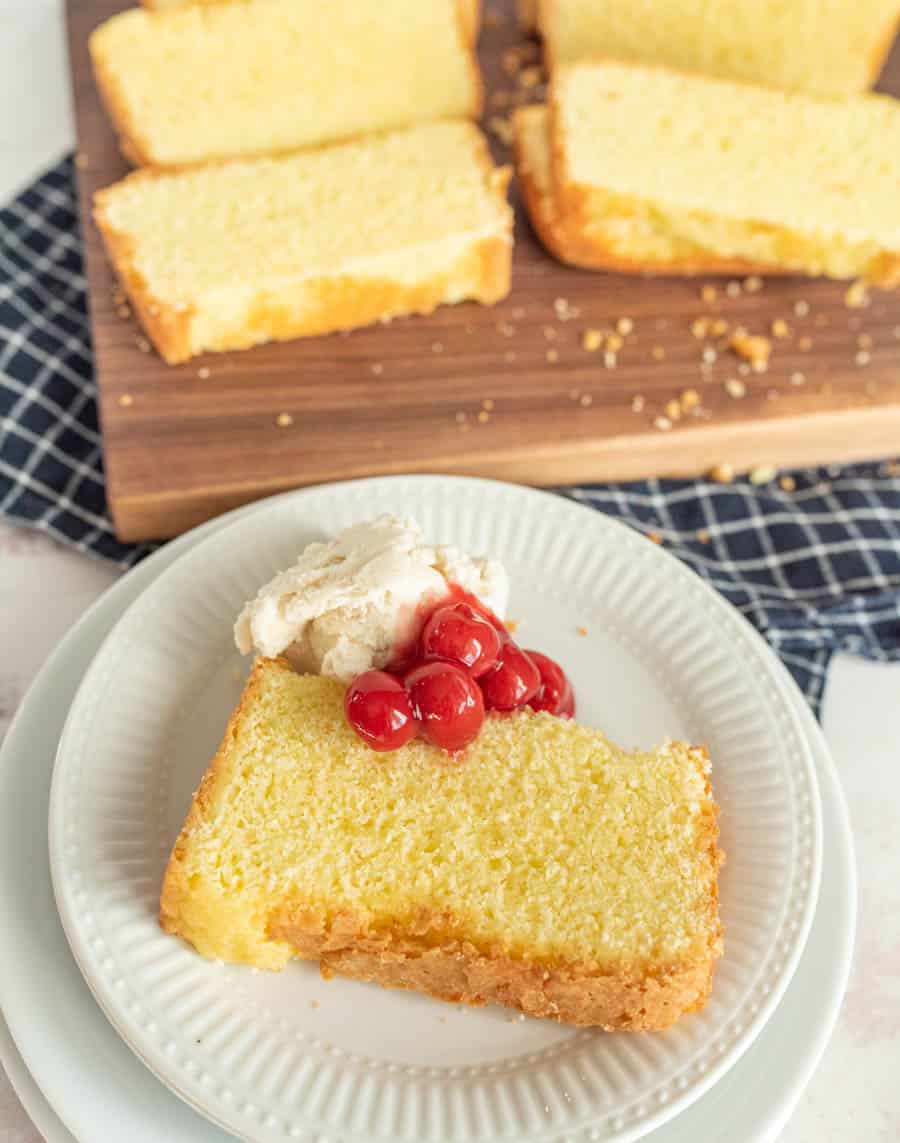 A plate of pound cake with ice cream and cherry topping and a cutting board with slices of pound cake in the background.
