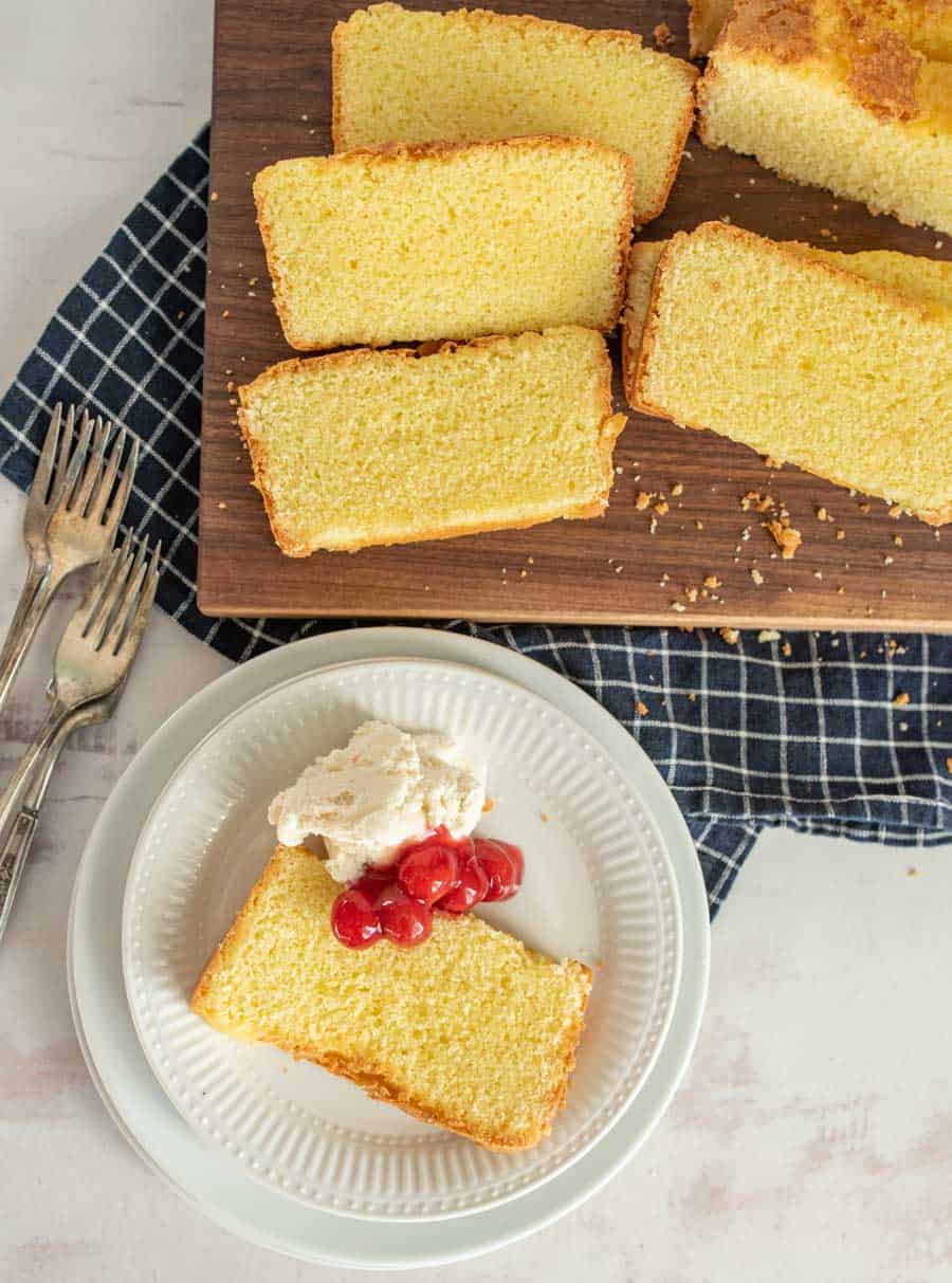 A top view of a slice of pound cake with ice cream and cherry topping and a cutting board of slices of pound cake.