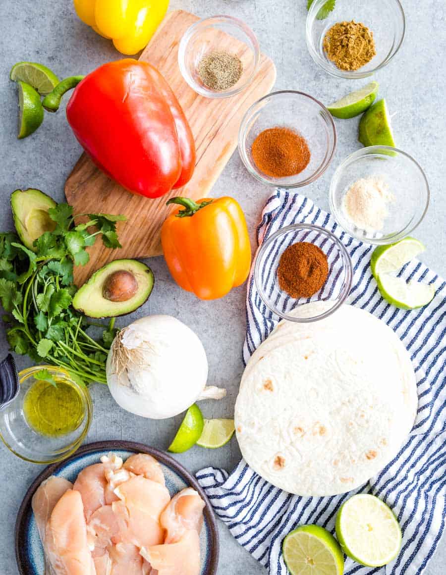 Colorful ingredients for sheet pan chicken fajitas, including bell peppers, tortillas, spices in small bowls, raw chicken, onion, avocado, lime wedges, cilantro, and olive oil, arranged on a striped cloth.