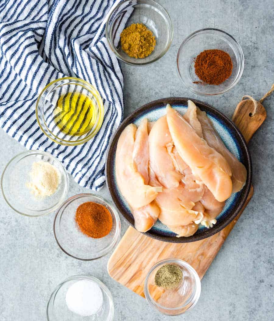 Raw chicken breast strips on a blue plate sit on a wooden board, surrounded by bowls of spices and oil—perfect for making sheet pan chicken fajitas. A striped cloth rests nearby on the gray surface.