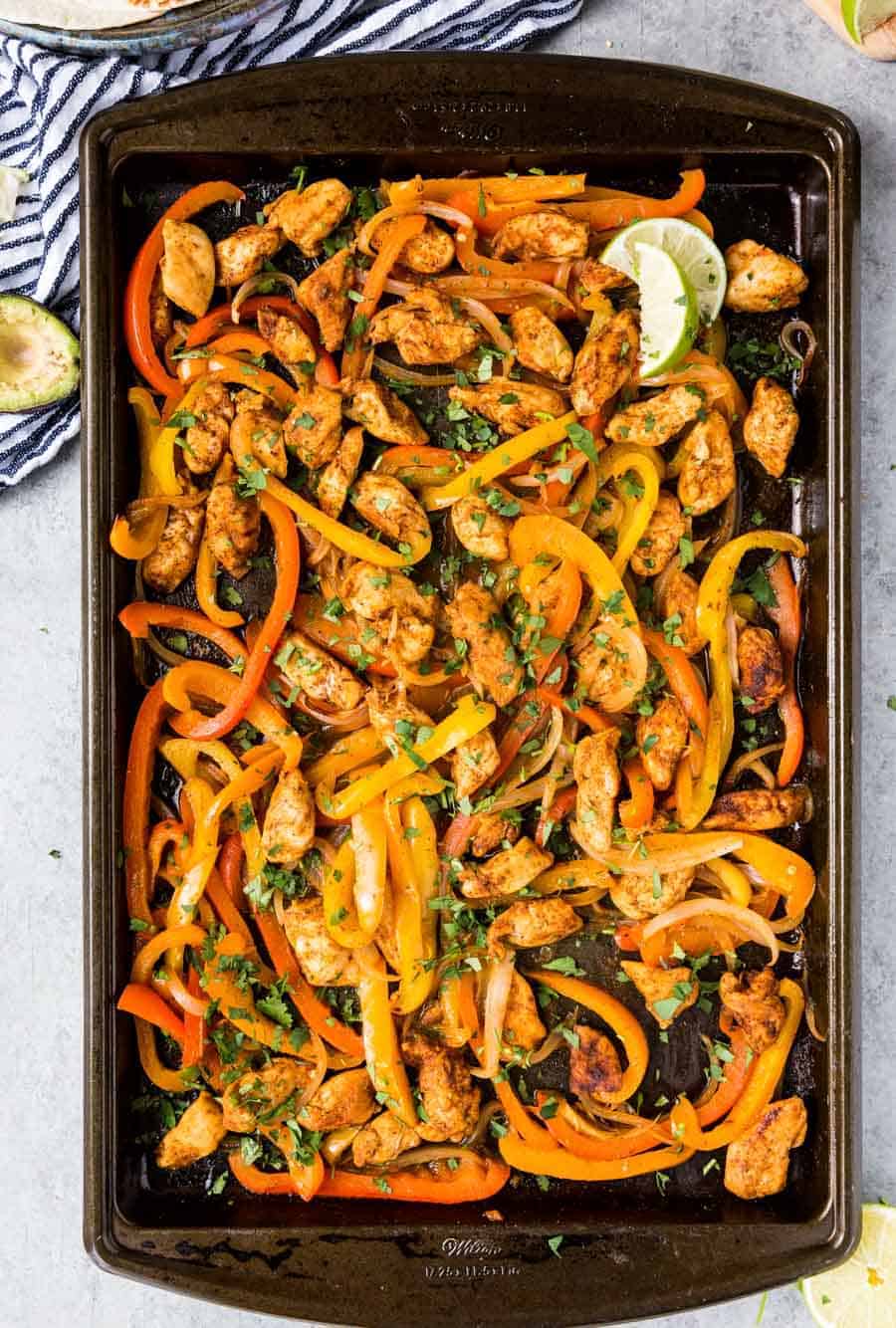 A baking sheet filled with cooked sheet pan chicken fajitas—juicy strips, red and yellow bell peppers, onions, all garnished with chopped herbs and lime wedges.