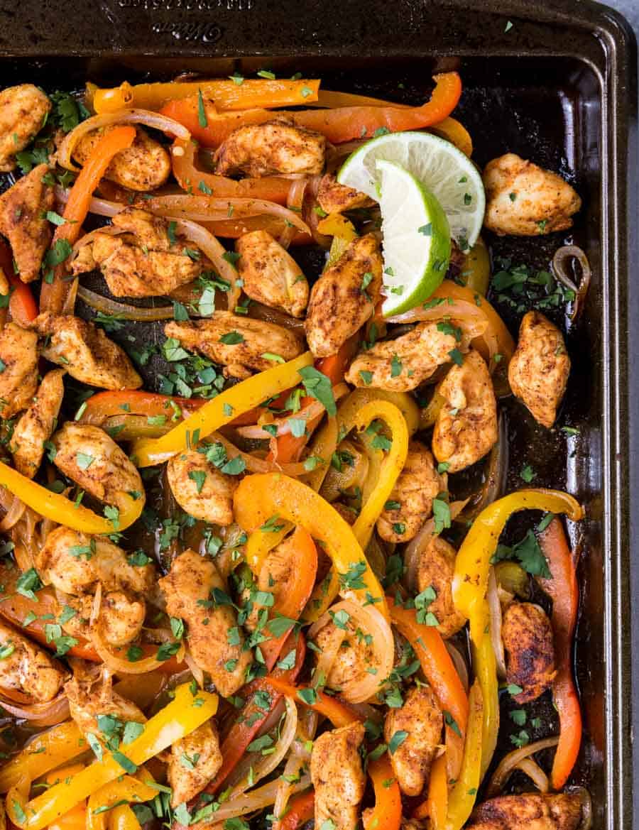 A baking sheet filled with cooked sheet pan chicken fajitas—sliced yellow and red bell peppers, onions, fresh cilantro, and a lime wedge—all perfectly seasoned and roasted.