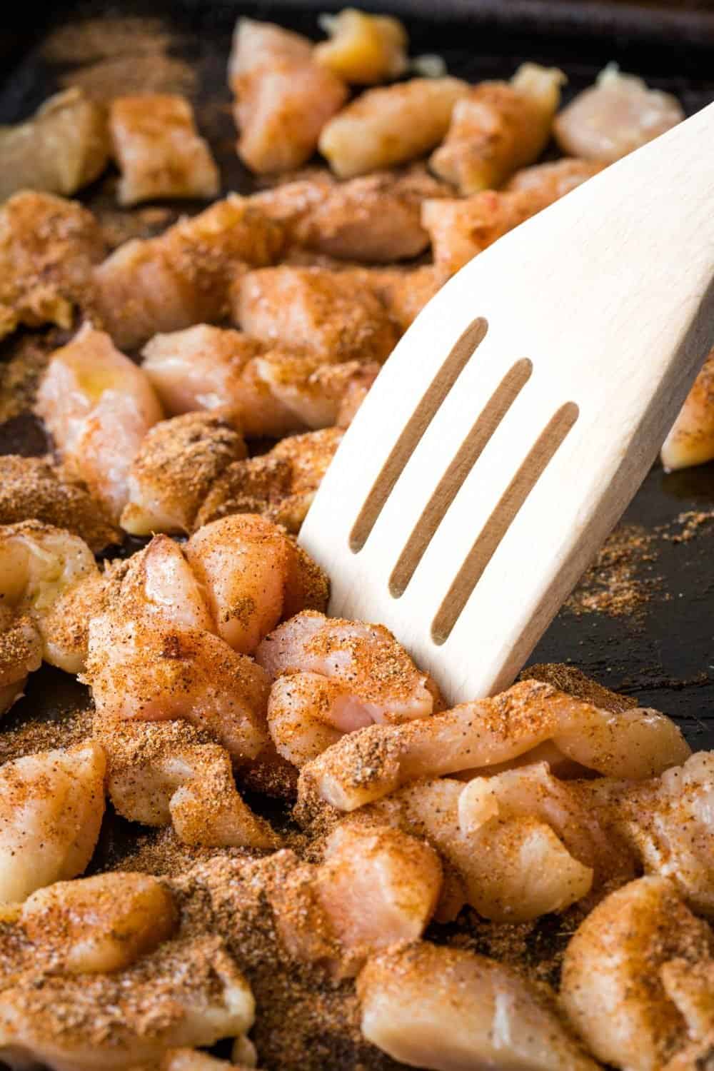 Seasoned raw chicken pieces being stirred on a pan with a wooden spatula, perfect for making flavorful sheet pan chicken fajitas.