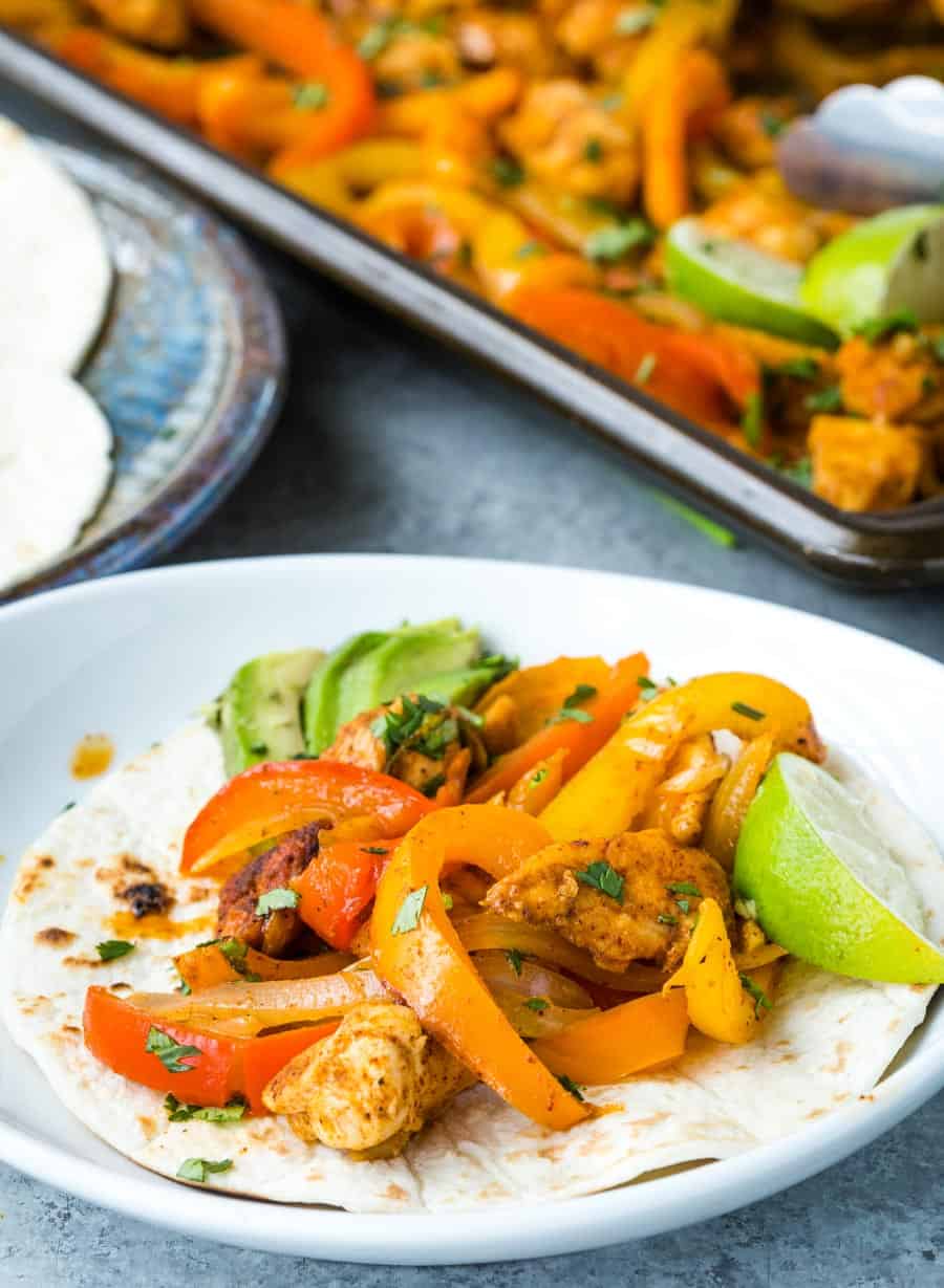 A tortilla topped with sheet pan chicken fajitas, sliced red and yellow bell peppers, avocado, and a lime wedge sits on a white plate. A tray with more fajita ingredients and tortillas is visible in the background.