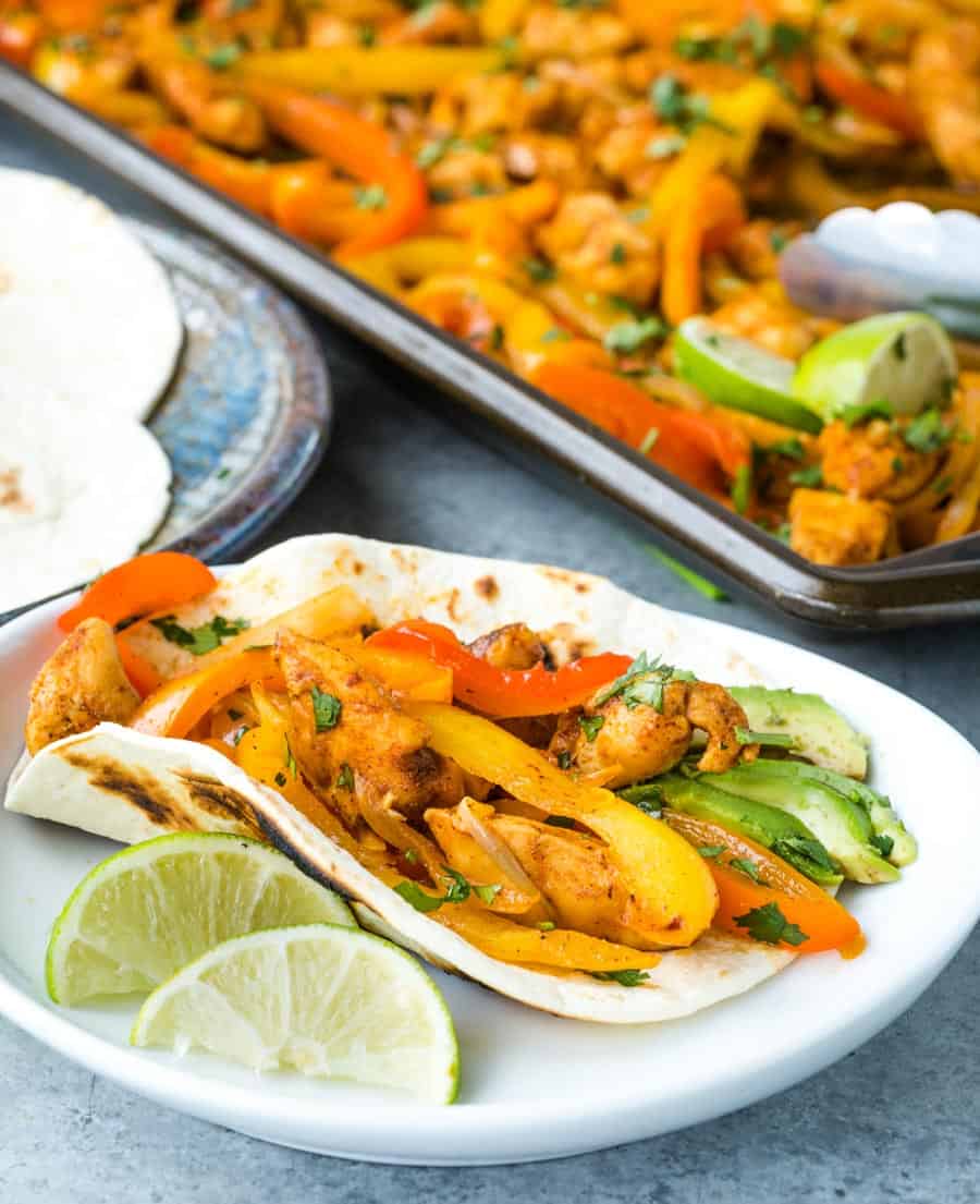 A taco filled with grilled chicken, yellow and red bell peppers, and avocado slices is served on a white plate with lime wedges. In the background, a tray of sheet pan chicken fajitas and tortillas is visible.