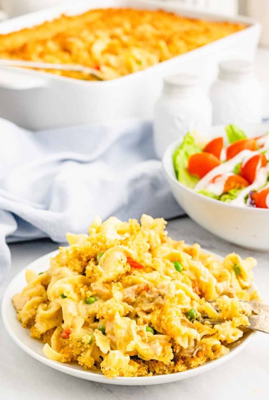 A close up plate of tuna noodle casserole with a baking dish of the casserole and side salad in the background.
