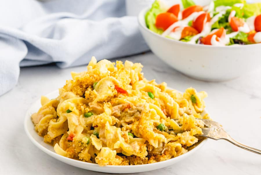 A close up picture of a plate of tuna noodle casserole with a fork in it and a bowl of salad in the background.