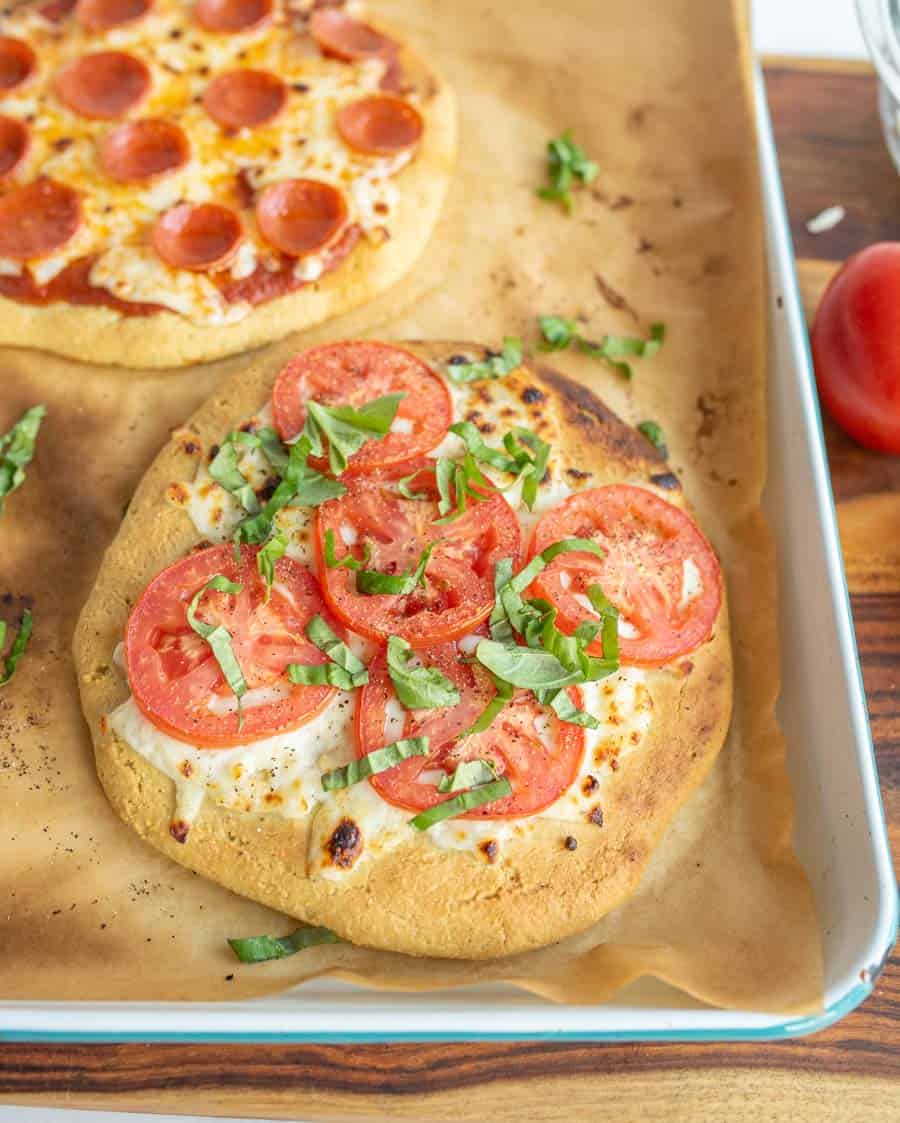 Two mini pizzas on a baking sheet, one with tomatoes and basil, the other with cheese and pepperoni.