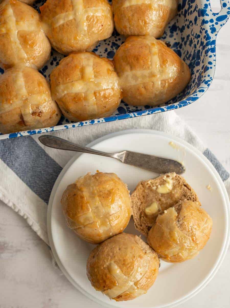 A top view of a plate with two hot cross buns on them, one cut in half with butter on it next to the rest of the pan of buns.