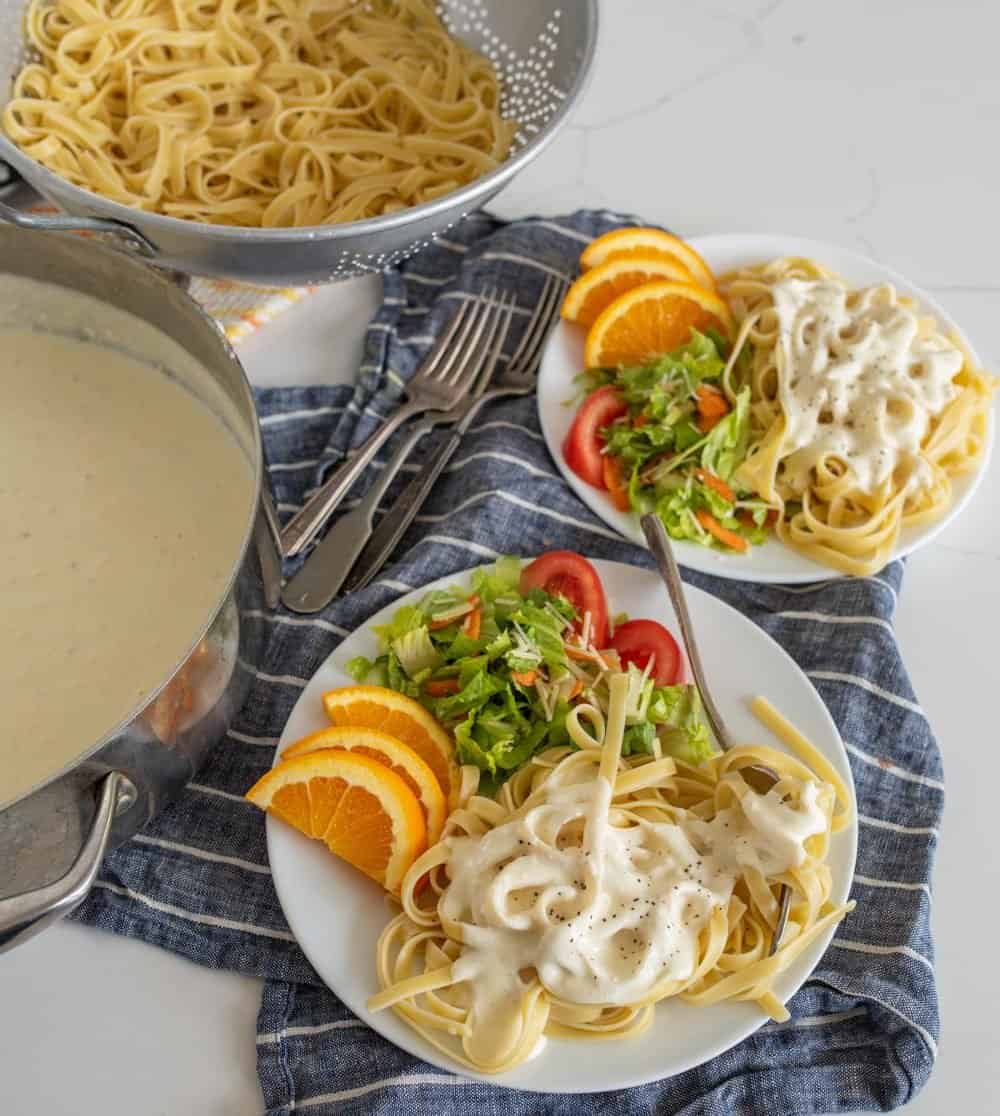 Plates of alfredo with salad and orange slices next to the pot of sauce and a colander of noodles.