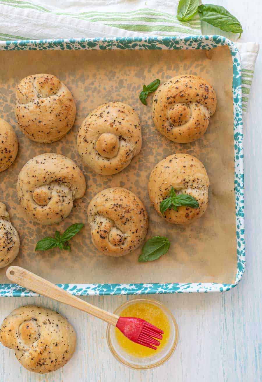 top view of garlic knots on a baking sheet with a bowl of melted butter next to it.