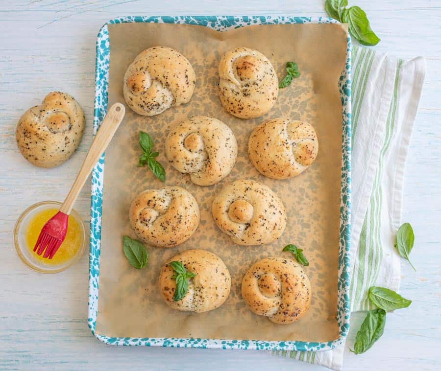 top view of garlic knots on a baking sheet with a bowl of melted butter next to it.