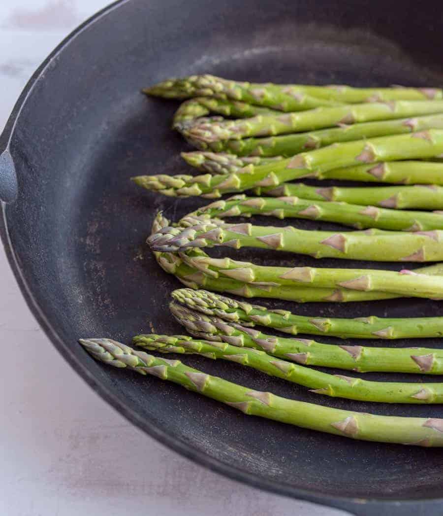 A bunch of fresh green asparagus spears, perfectly sautéed, arranged in a black cast iron pan on a light, textured surface.