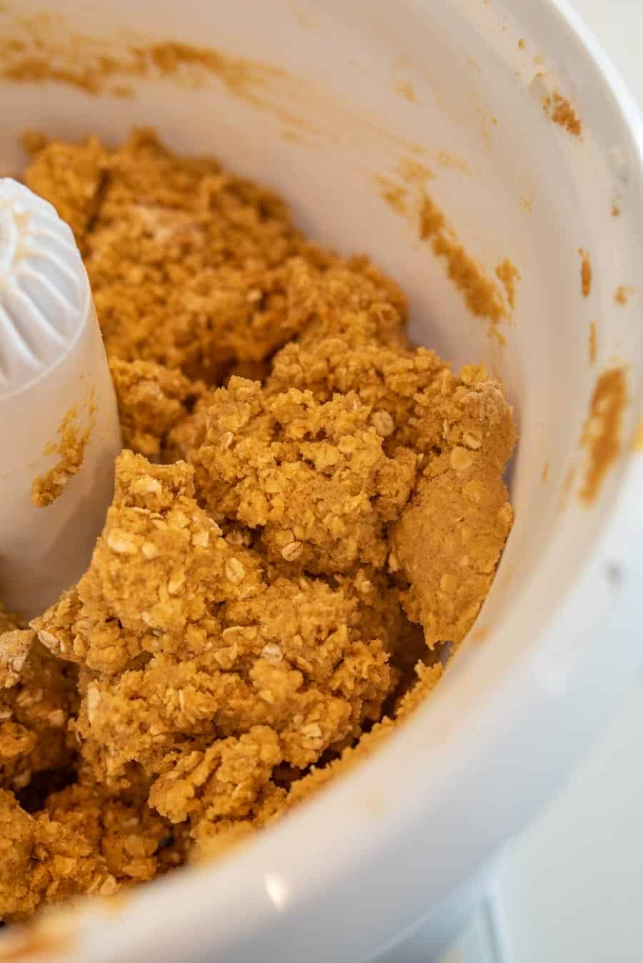 Close-up of oatmeal cookie dough being mixed in a white food processor, showing a crumbly, golden-brown texture with visible oats.