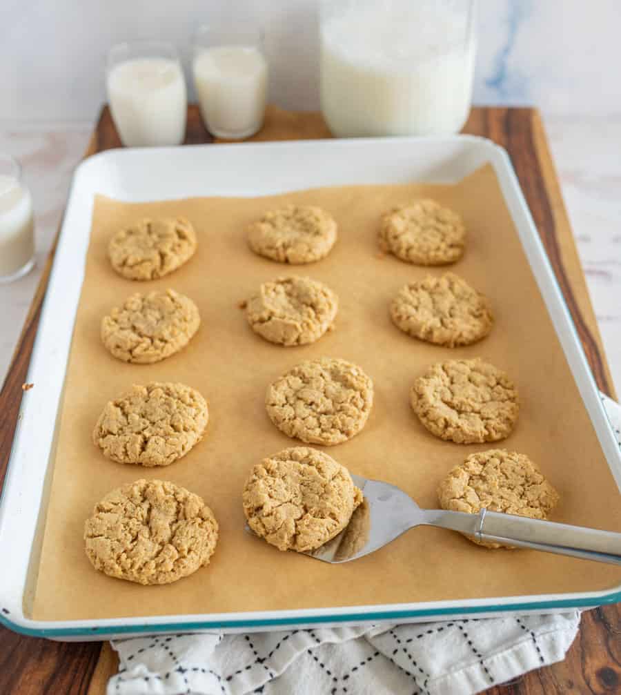A baking tray lined with parchment paper holds twelve freshly baked oatmeal cookies, with a metal spatula lifting one. Glasses and a jar of milk are seen in the background on a wooden surface.