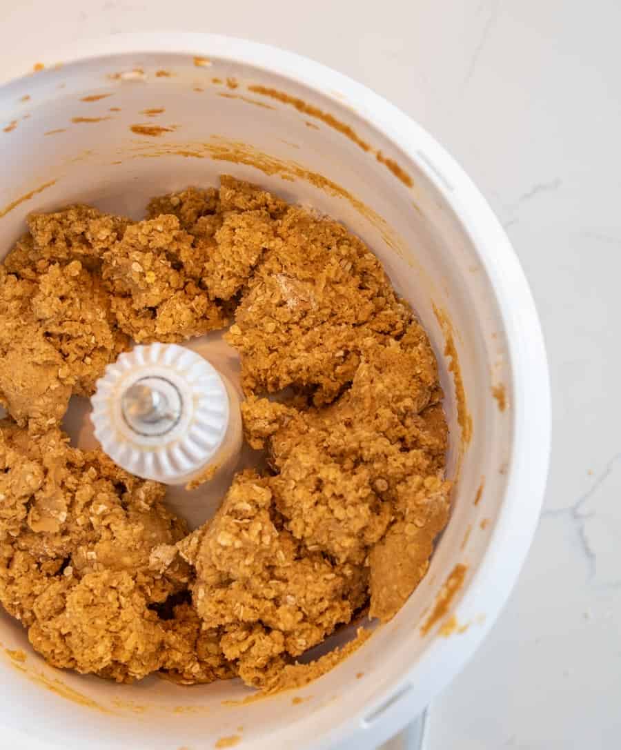 A food processor bowl containing a thick, light brown dough mixture pressed against the sides, viewed from above on a white countertop.