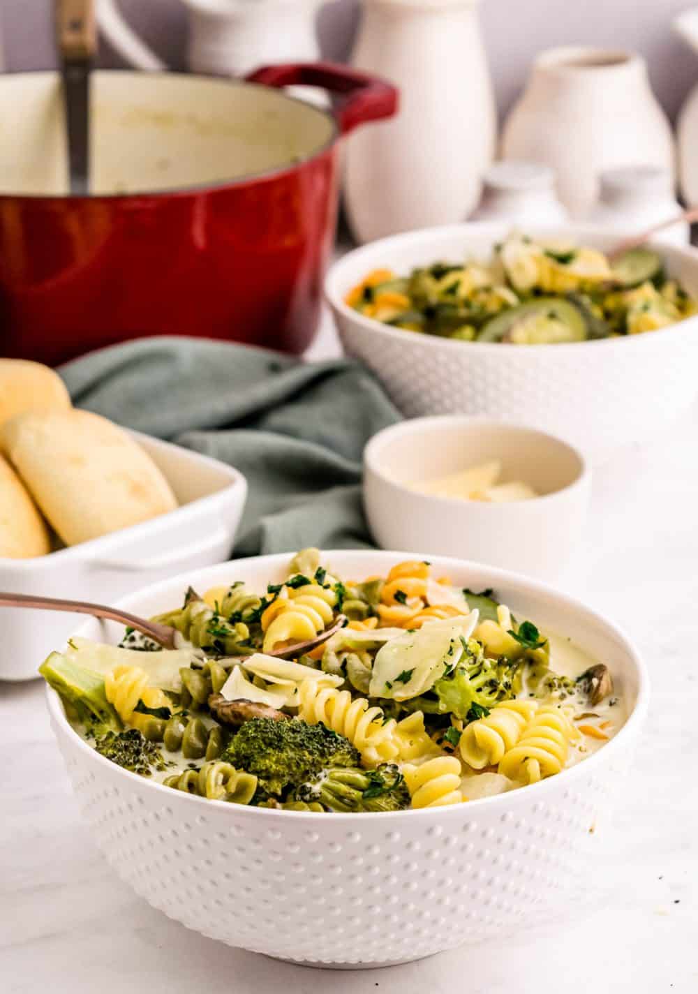 A bowl of pasta primavera with another bowl and pot in the background.