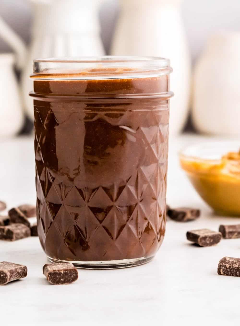 Side view of jar of peanut butter hot fudge with chocolate chunks around it.