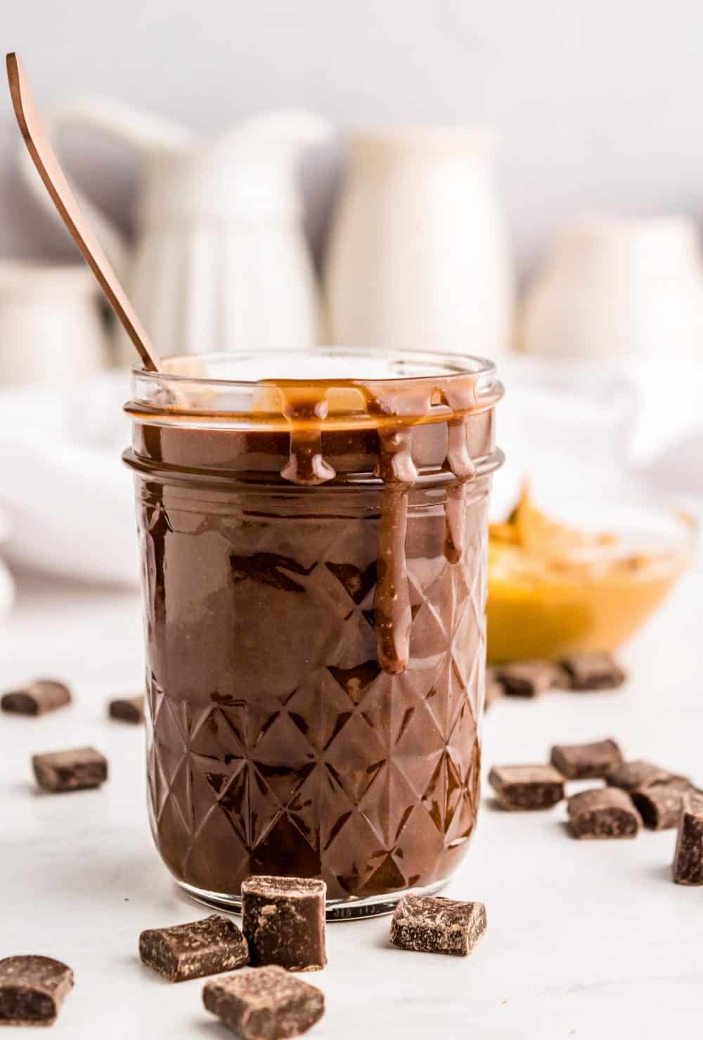 A jar of peanut butter hot fudge, some dripping down the side, with chocolate chunks around it.
