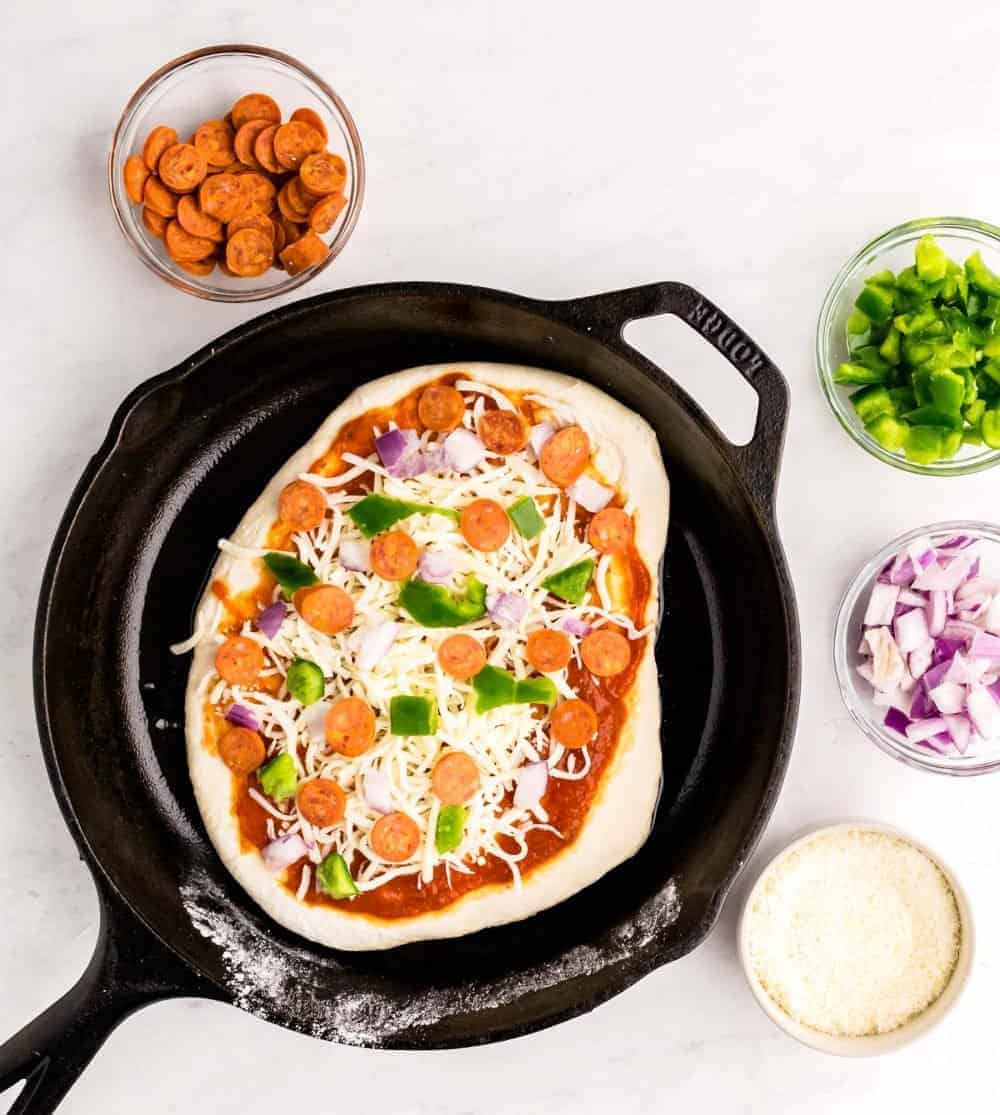 A top view of uncooked pizza in a skillet with ingredients in bowls surrounding the skillet.