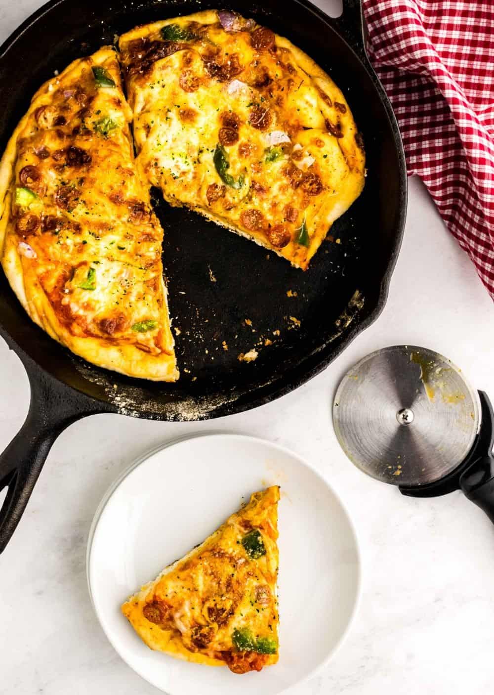 A top view of a slice of skillet pizza on a white plate with the rest of the pizza in the skillet, next to it.